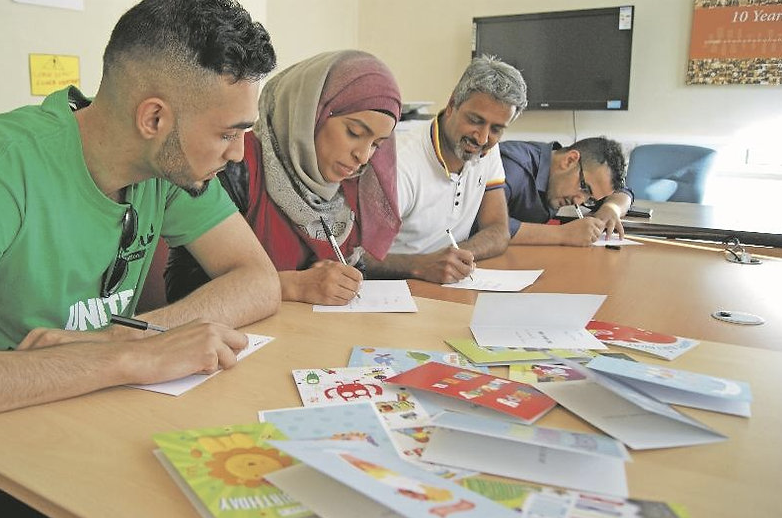 Four adults sat at a table indoors writing on blank paper with a pile of cards in front of them.